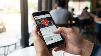 Primer plano de una persona sosteniendo un smartphone con la aplicación de YouTube abierta, mostrando el logotipo rojo y el botón de reproducción.