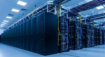 Vista de un centro de datos con largas filas de gabinetes de servidores negros, cableado complejo azul y amarillo sobre los racks y un techo iluminado.