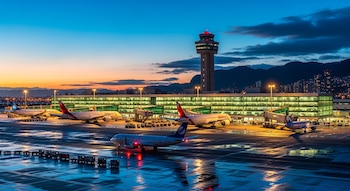 Aeropuerto El Dorado en Bogotá al atardecer, con aviones estacionados en la plataforma mojada, torre de control y terminal iluminados, y montañas al fondo.