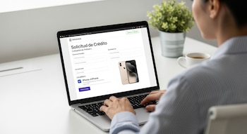 Vista por encima del hombro de una persona adulta usando una laptop en un escritorio blanco, mostrando un formulario de solicitud de crédito con un iPhone como garantía.