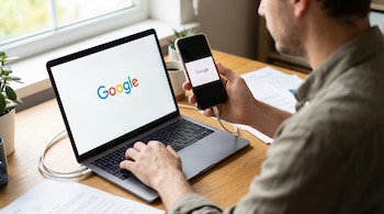 Un hombre sentado en un escritorio de madera opera un laptop y un teléfono móvil. Ambas pantallas muestran el logo de Google. Cables conectan los dispositivos.