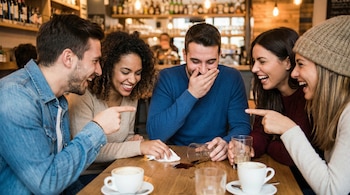 Cinco personas jóvenes ríen alrededor de una mesa de madera en una cafetería. Un vaso volcado ha derramado una bebida oscura en la mesa, y hay tazas de café.