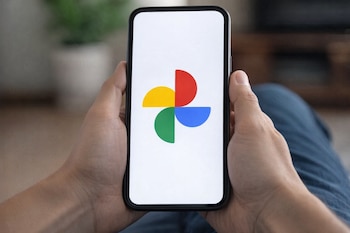 Primer plano de dos manos sosteniendo un smartphone con el logo de Google Fotos en la pantalla brillante, contra un fondo ligeramente desenfocado.