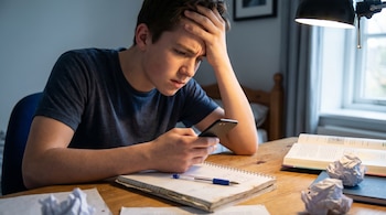 Primer plano de un adolescente con camiseta azul, estresado, sosteniendo un teléfono. Está en un escritorio con un cuaderno abierto, una lapicera y papeles arrugados.