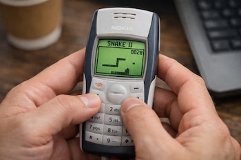 Manos sosteniendo un teléfono Nokia 1100 en primer plano, con el juego Snake II visible en su pantalla monocromática verde.